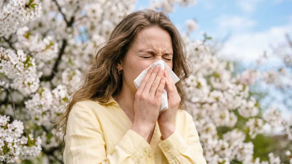 young woman sneezing in spring garden with blooming trees, experiencing seasonal allergies